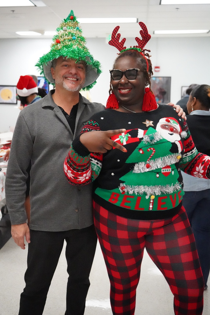 A man with a Christmas tree hat and a woman wearing reindeer antlers and a holiday sweater stand together for a photo in a building and smile. The woman is doing the peace sign with her hand.