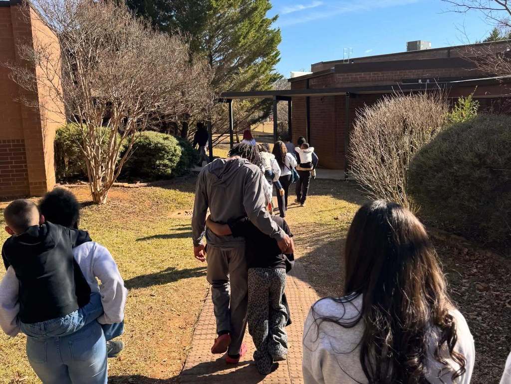 Clay-Chalkville High School students and Clay Elementary students walk together outside a school building. One pair of students hug and another student carries a younger student on their back.