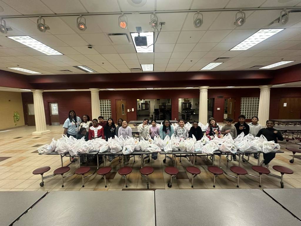 Brighton School Peer Helpers stand together for a photo in a lunchroom. They stand behind lunch room tables with bags on them.