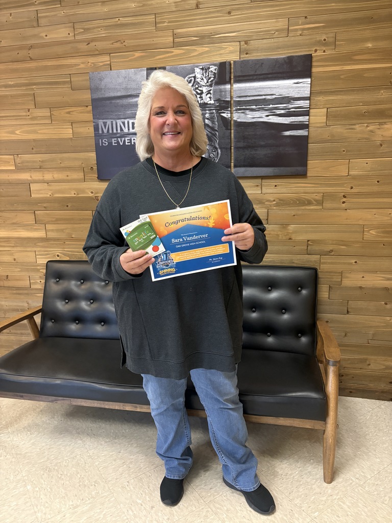 Ms. Sara Vanderver smiles and holds a JEFCOED Strong Award in her hand while standing inside a building. She stands in front of a small couch and a motivational poster of a kitten looking into a puddle and seeing a tiger. Text on the poster says "Mindset is Everything."