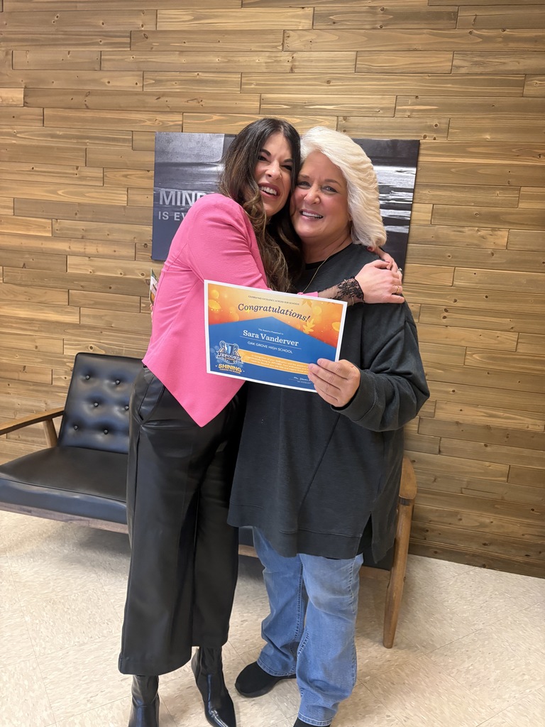 Sara Vanderver holds a JEFCOED Strong Award in her hand as she hugs Oak Grove High School Assistant Principal, Kristin Pederson. The two stand in a building in front of a wall, couch and motivational poster.