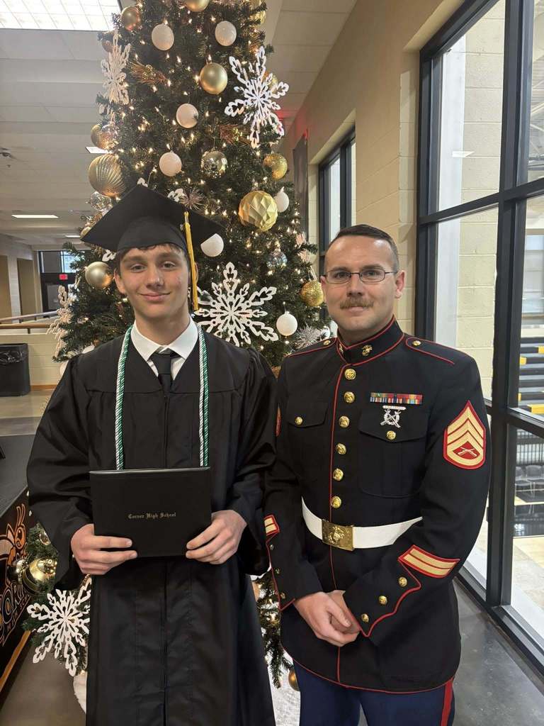 A student wearing a graduation cap and gown and a man in a Marine uniform stand together for a photo inside a school hallway.