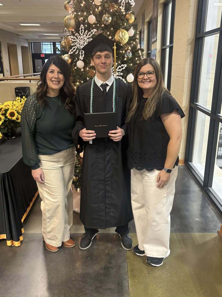 Two women and a student in a graduation cap and gown stand together for a photo inside a school hallway.