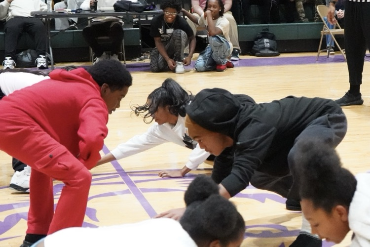 Students in a school gym squat down in the middle of the basketball court and face each other. One of the students is reaching for something on the ground.