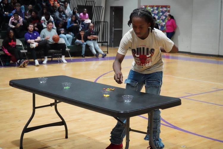 A student stands at a table in a school gym and places skittles in a cup.