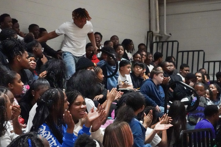 Students sitting in bleachers in a school gym cheer for a fellow student as he runs down the bleachers.