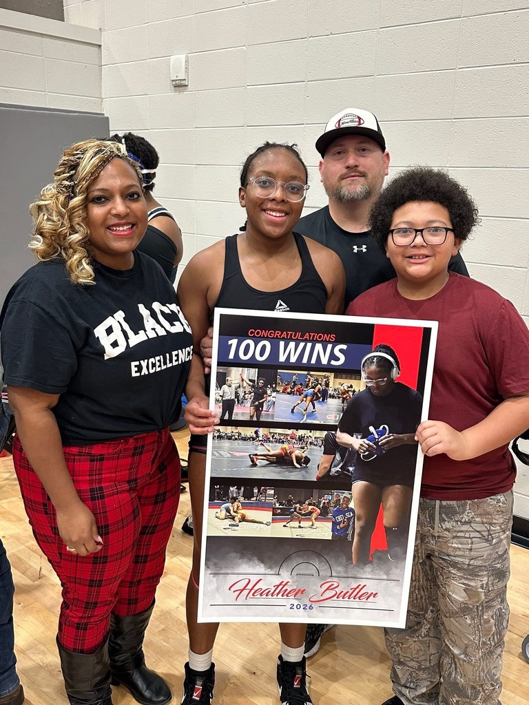 A woman, a high school student, a man, and a boy stand together for a photo in a gymnasium. The high school student and boy hold up a poster with images of the high school student and text that says "Congratulations 100 wins Heather Butler 2026."