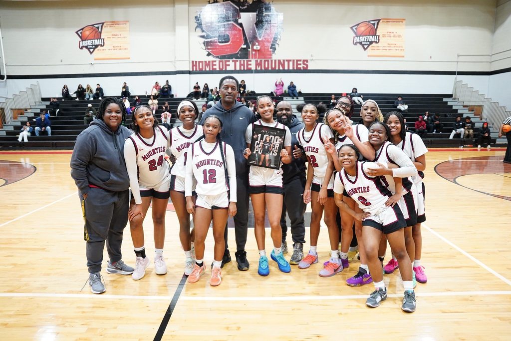Shades Valley High School girls basketball players stand together in uniform on the basketball court for a photo with three adults. Player Zoey Little stands  in the middle and holds up a framed graphic announcing her reaching 1,000 career basketball points.