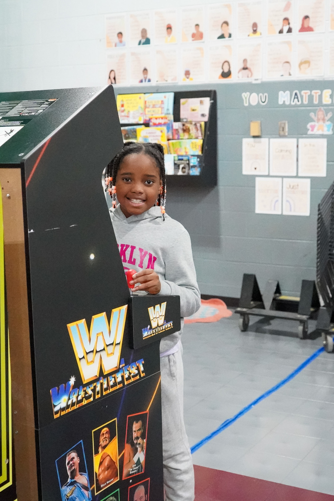 A girl smiles while playing an arcade-style video game inside a school gym.
