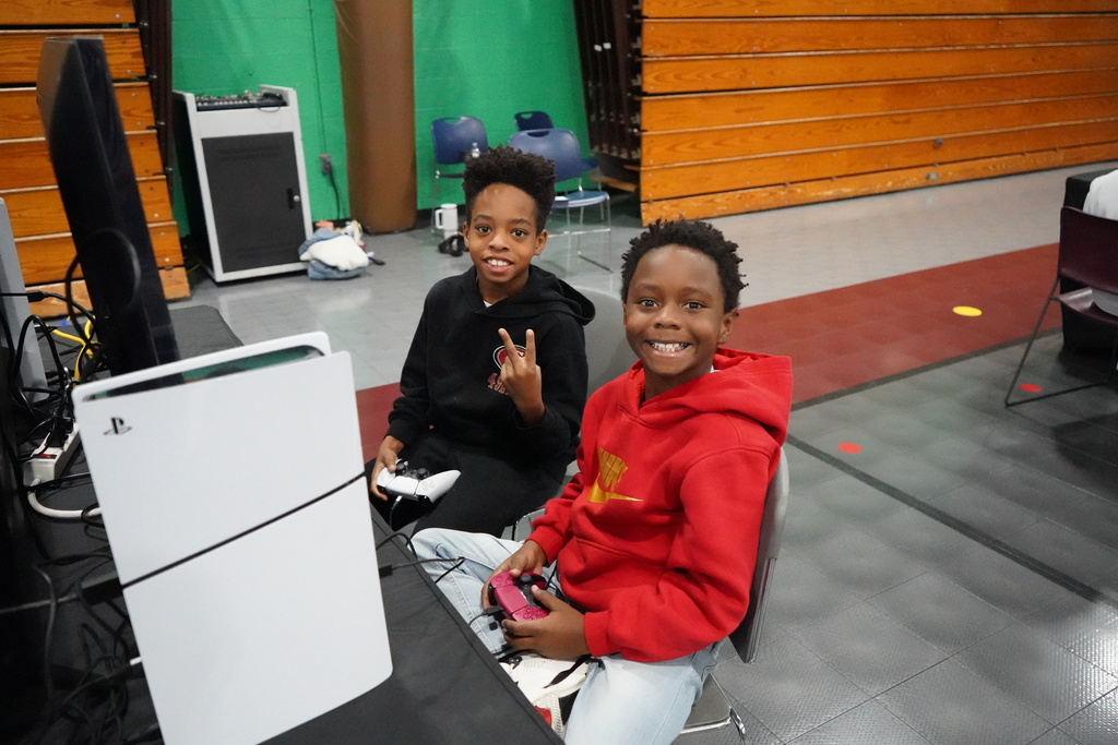 Two boys smile for a photo while playing a video game inside a school gym. One of the boys holds up a peace sign with his hands.