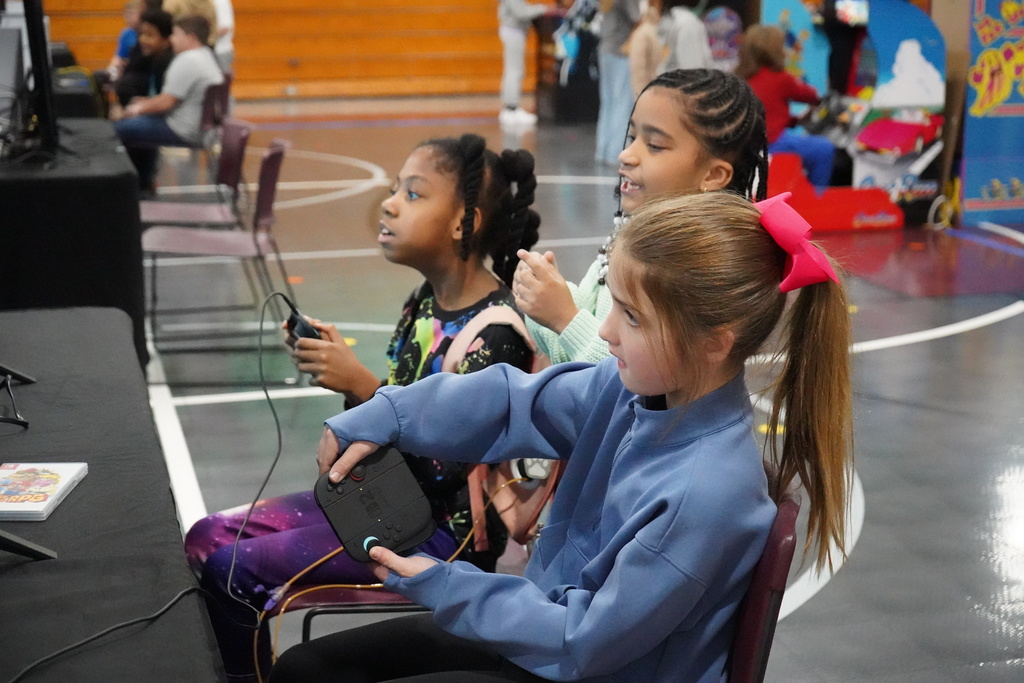 Two girls sit at a table and play video games inside a school gym. Another girl stands between them and talks.