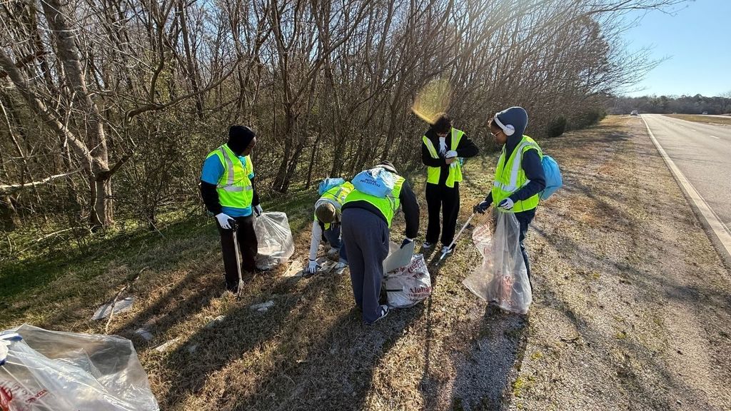 A group of people wearing yellow work vests pick up trash off the side of the road.