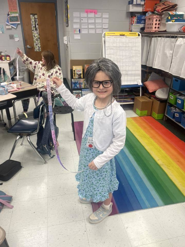 A student dressed like an older person stands in a classroom and smiles for a photo.