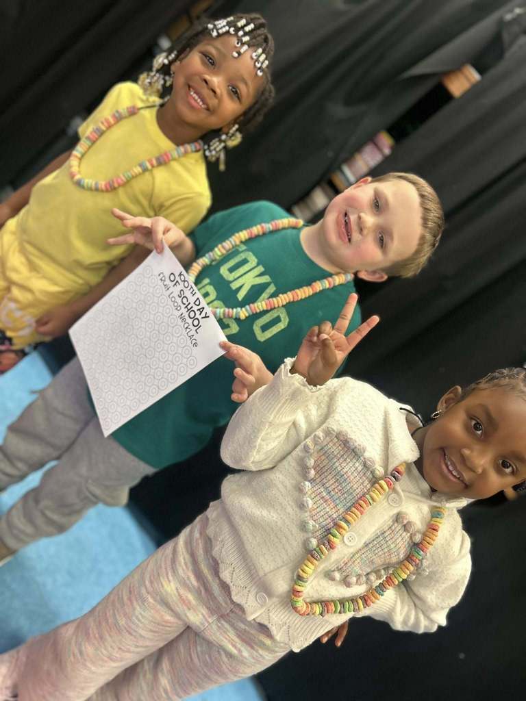 Elementary students stand for a photo and smile. Each of the students wears a necklace made of fruit loops and the student in the middle holds a paper that says "100th day of school fruit loop necklace."