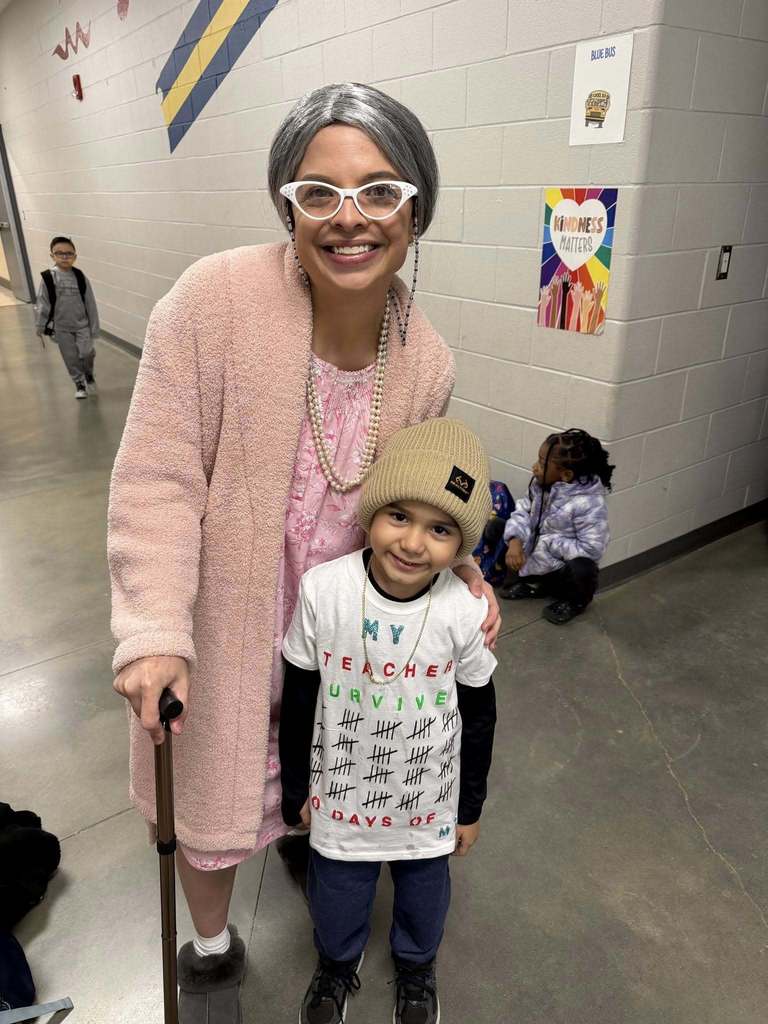 A teacher dressed like an older person poses next to a student in a school hallway.