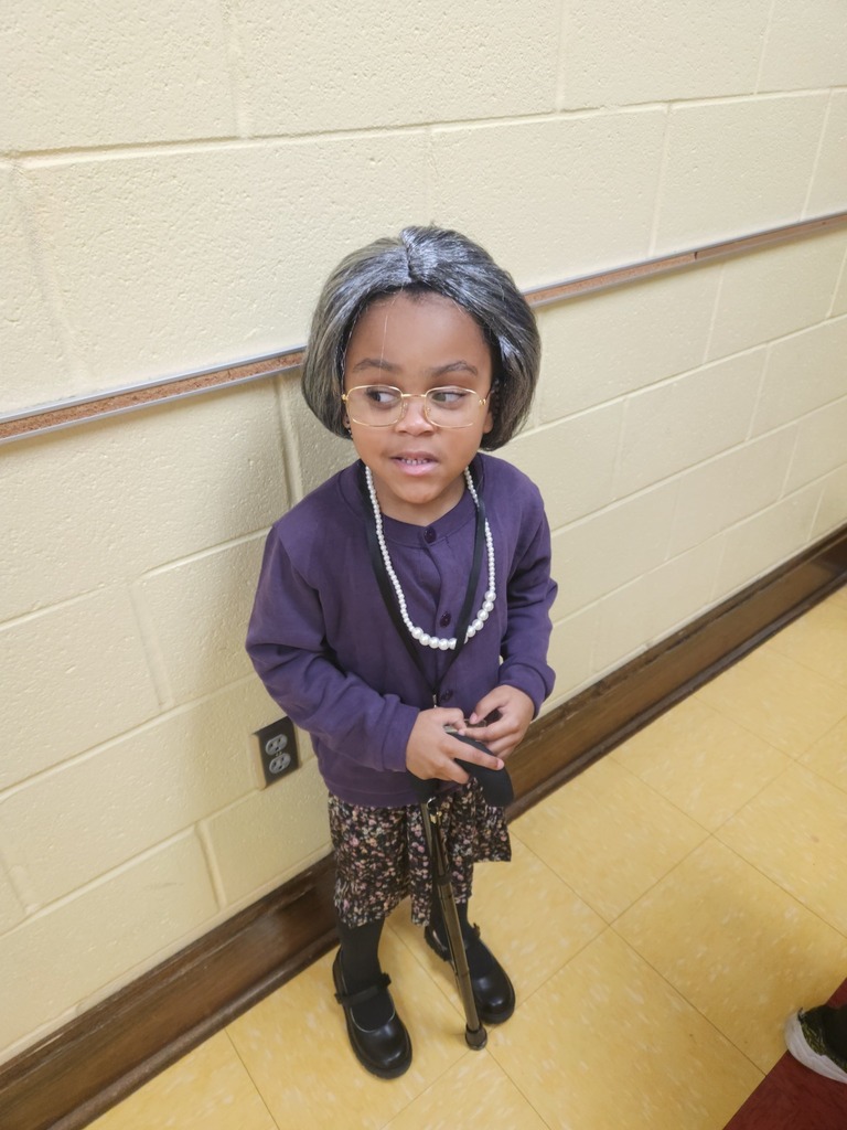 An elementary school student stands for a photo in a school hallway. The student is dressed like an older person.