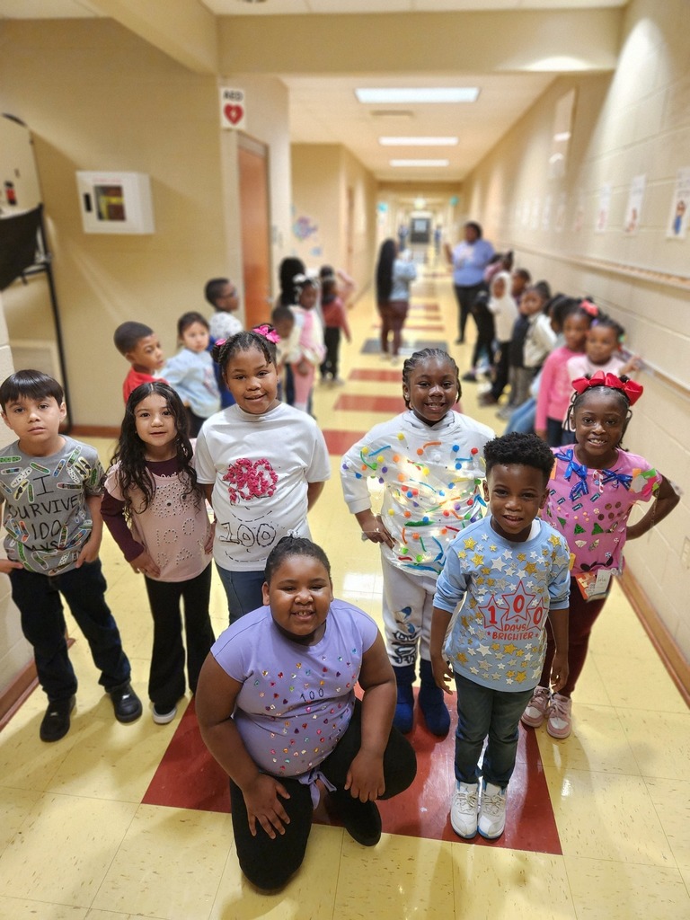 Students stand for a photo in a school hallway. They wear hand-decorated shirts themed for the 100th day of school.