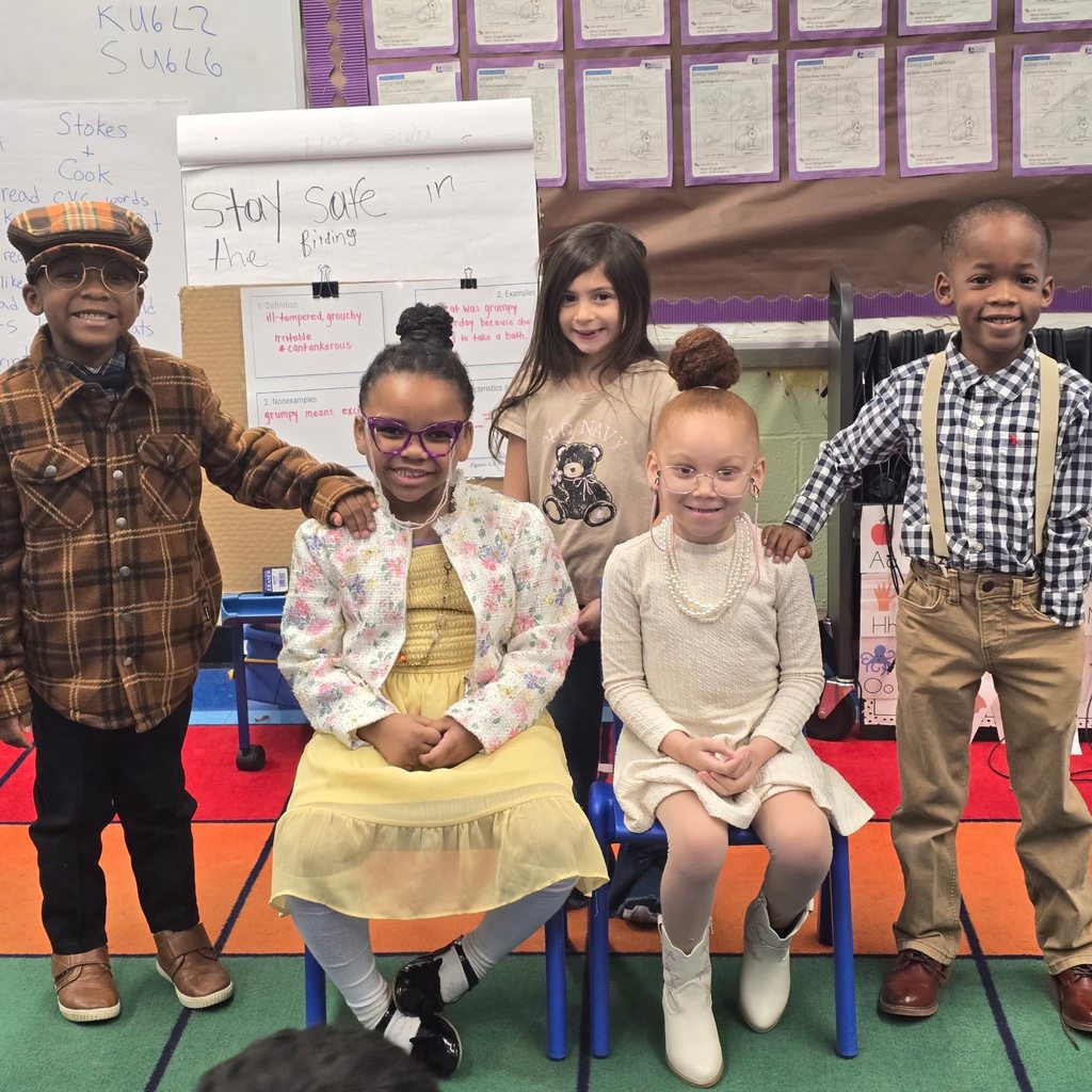 Elementary school students dressed like older people stand and sit for a photo inside a school classroom.