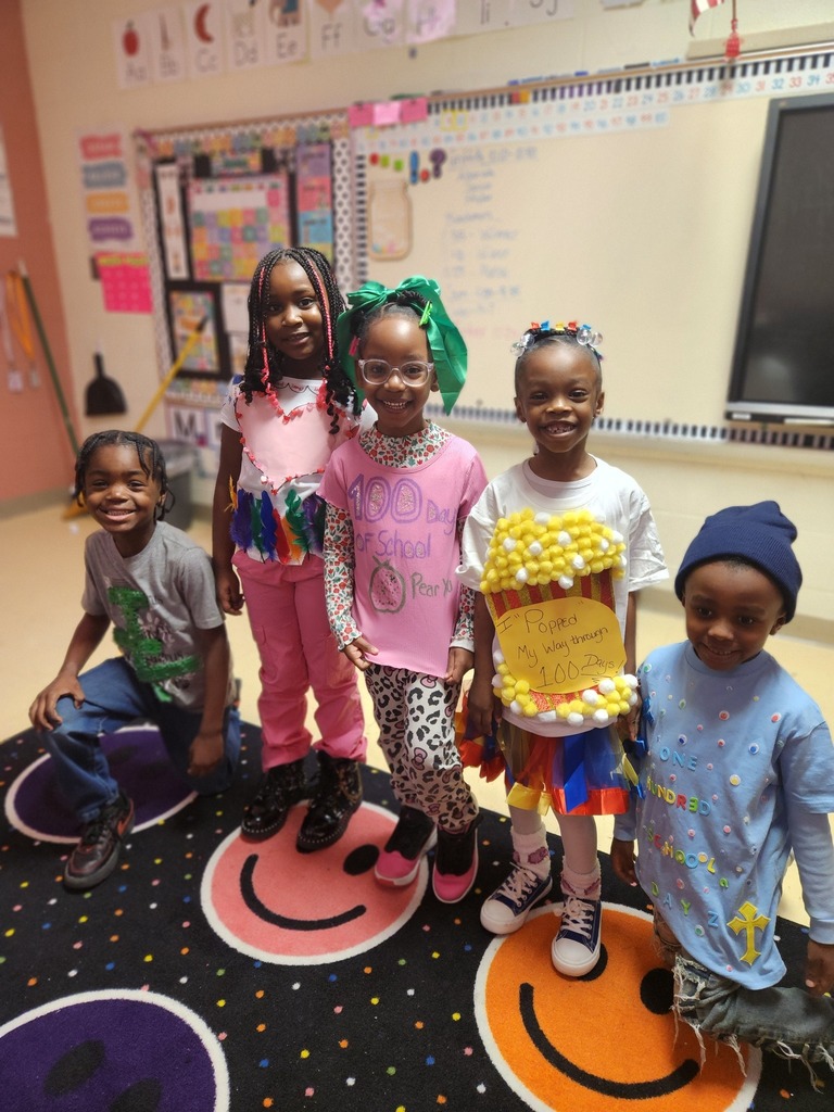 Elementary students stand for a photo inside a classroom. They wear shirts themed for the 100th day of school.