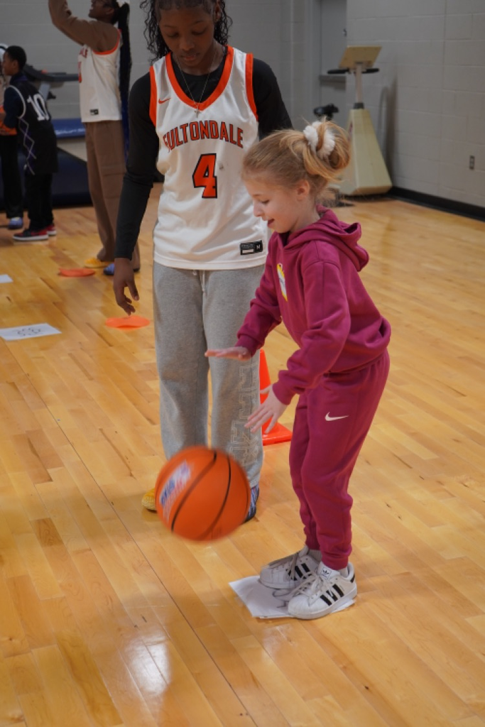 An elementary school aged girl dribbles a basketball inside a small gym. A Fultondale High School basketball player stands next to her.