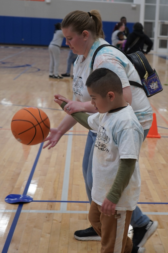 A teacher helps a student dribble a basketball inside a school gym.