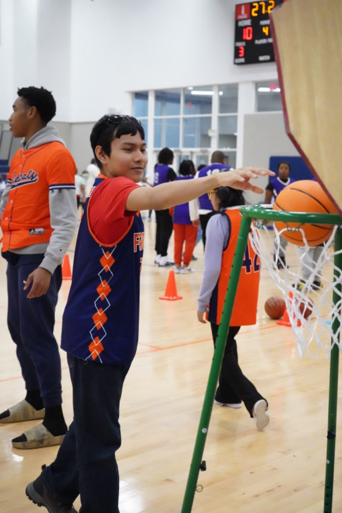 A student throws a basketball into a basketball hoop while standing inside a crowded school gym.