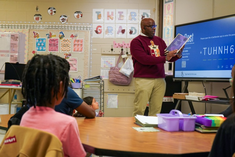 A man wearing a Kappa Alpha Psi shirt reads to a classroom of students.