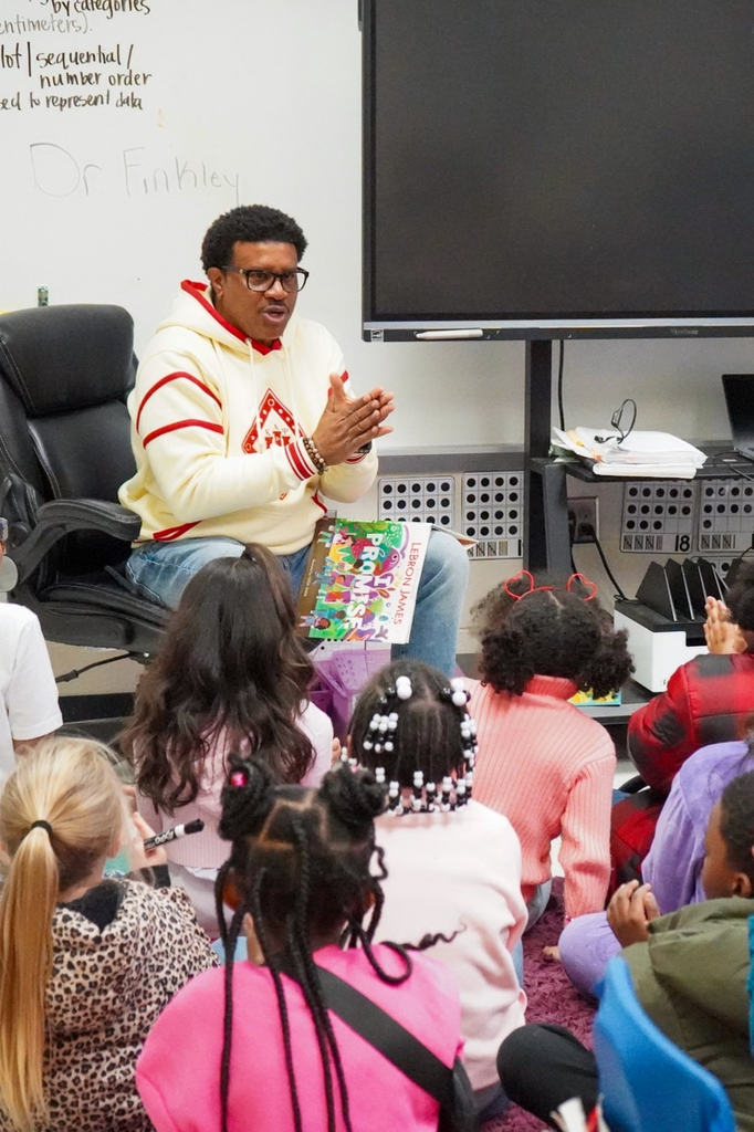 A man sits in a chair and reads a book to a classroom of students as they sit on the floor.