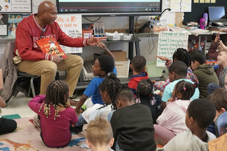 A man sits in a chair and reads to a classroom of students sitting on the floor.