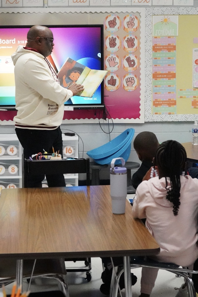 A man reads to students in a classroom as they sit at tables.