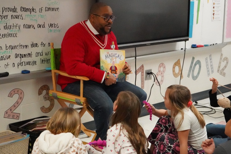 A man sits in a chair and reads to a group of students sitting in the floor of a classroom.