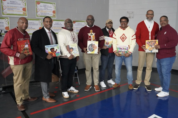 A group of Kappa Alpha Psi Fraternity members stand together for a photo in a school gym while holding books.