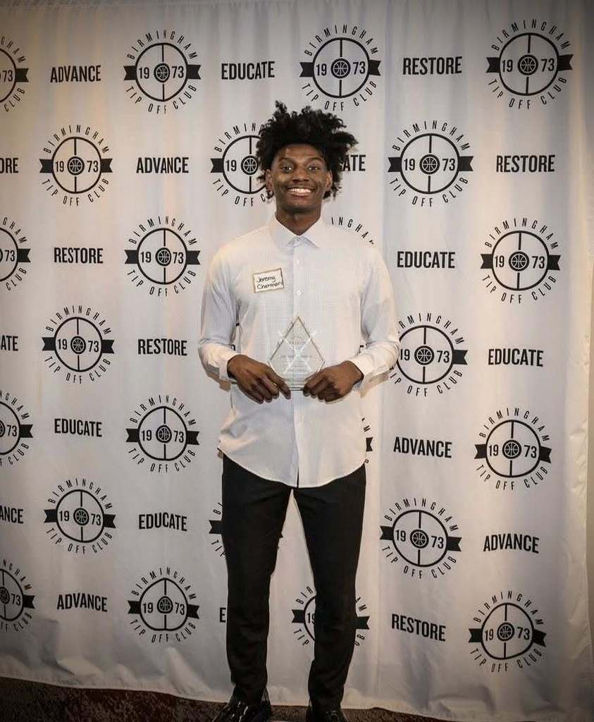 Shades Valley High School student Jeremy Chatman smiles and holds an award while standing in front of a Birmingham Tip Off Club photo backdrop.