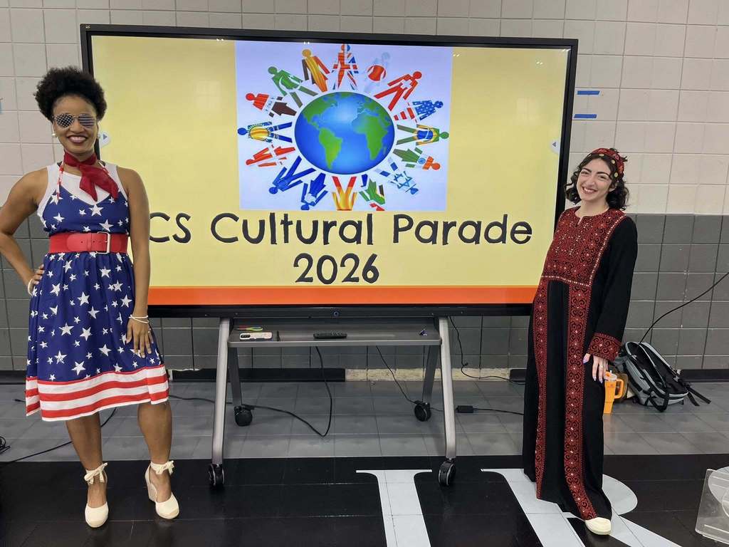 Two women stand together for a photo in front of a monitor that says "ICS Cultural Parade 2026." One woman wears a dress with stars and red and white stripes on it. Another woman wears a long black and red gown with a matching hair piece.