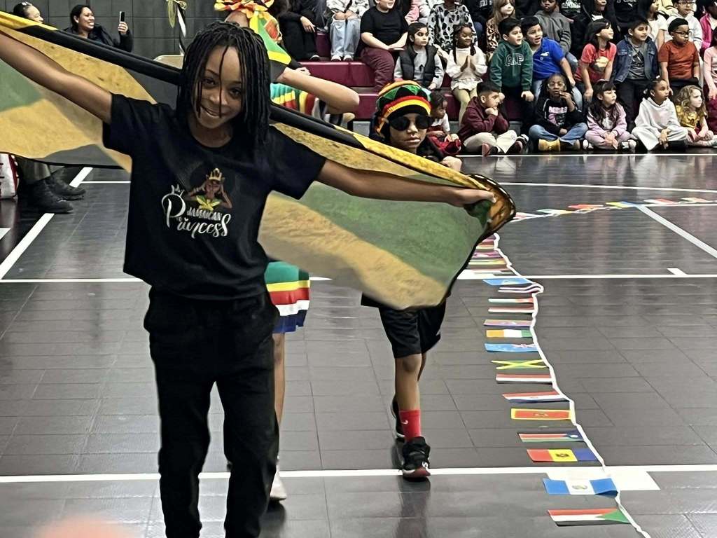 Students representing Jamaica at Irondale Community School's Cultural Parade walk through the school gym. One student holds a Jamaican flag and wears a shirt that says "Jamaican Princess."