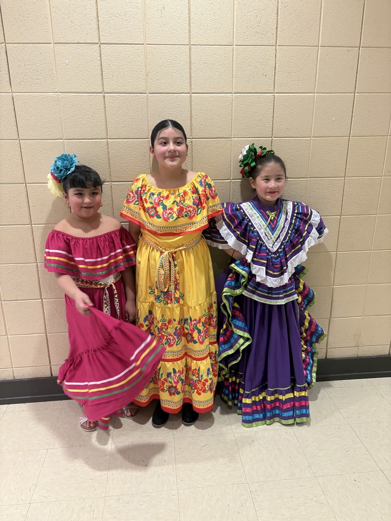 Three girls stand together in a school hallway wearing brightly colored dresses. Two of the girls wear flowers in their hair.