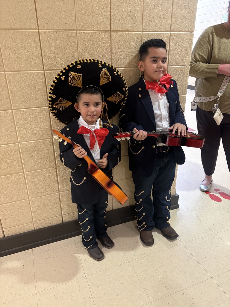Two students stand in the hallway of Irondale Community School for a photo. They both wear navy blue suits with buttons and chains embellished on the arms and pants. They also each hold a small guitar. One of the boys wears a Sombrero Charro.