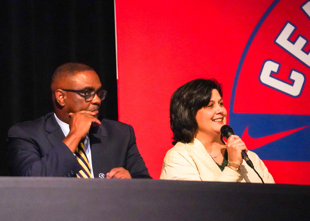 A man and a woman sit behind a table as part of a panel. The woman is holding a microphone. 