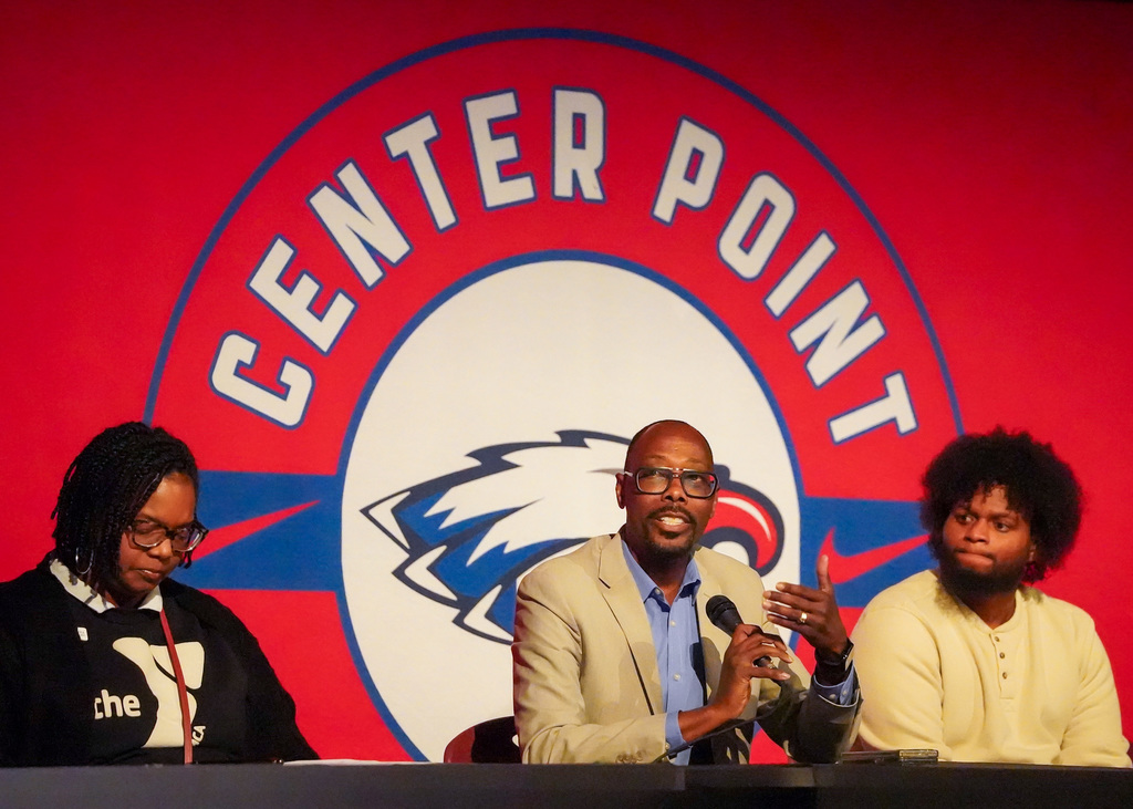Three people sit on a panel. A bright red Center Point High School banner is behind them. The man in the middle holds a microphone and is speaking. 