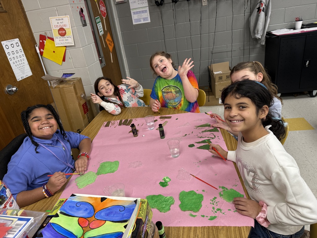  A group of young students sits at a school table. They are painting on a piece of pink paper. They are also smiling at the camera and making silly faces. 