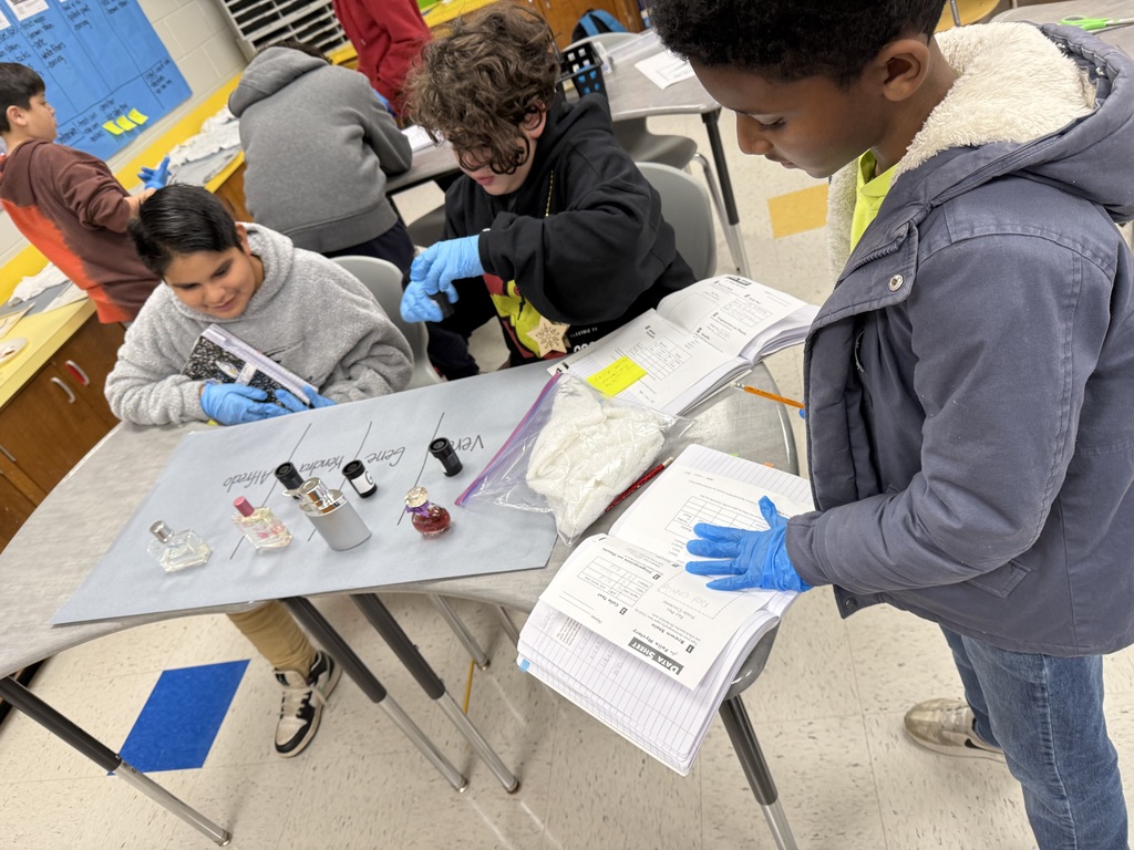 A group of elementary school students work on a project. they are wearing blue gloves, and are studying from a book and observing bottles of liquid.