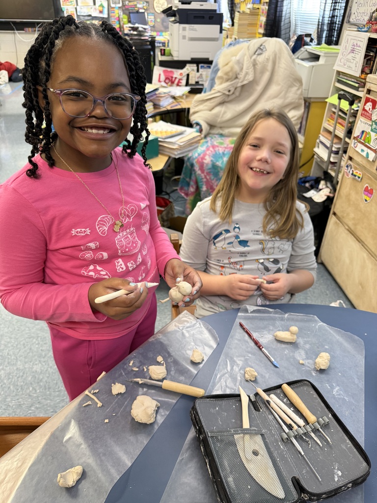 Two female elementary school students are working with clay and tools. They are smiling at the camera. 
