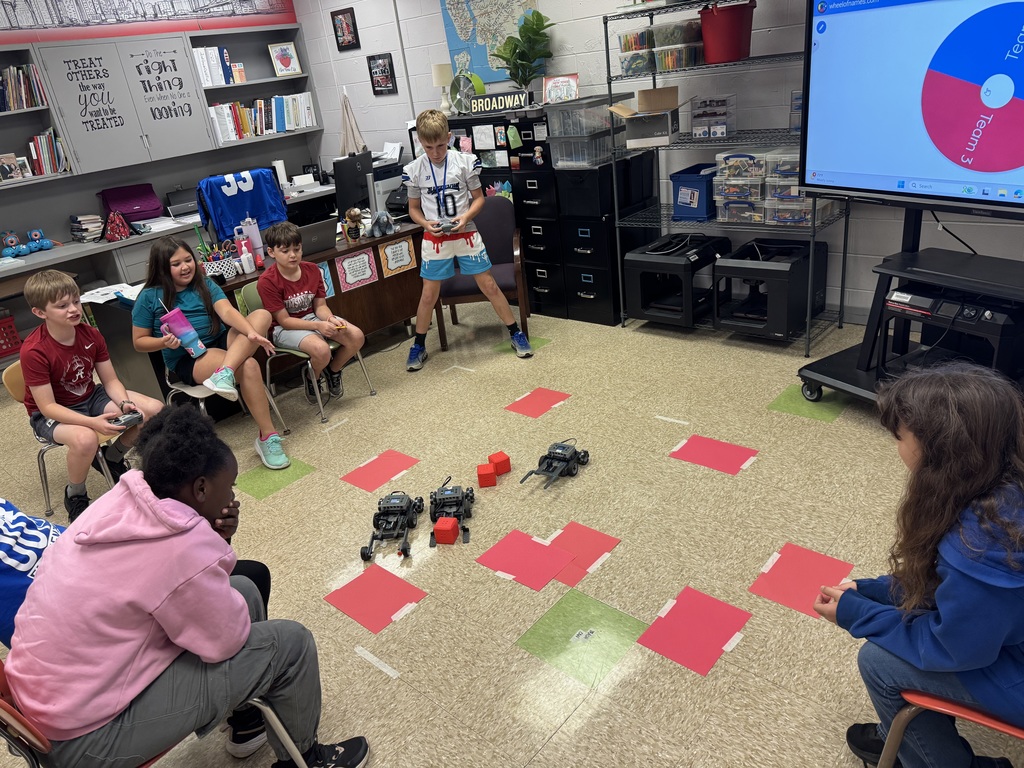 A group of elementary school students work to move robots through a maze on the floor. 