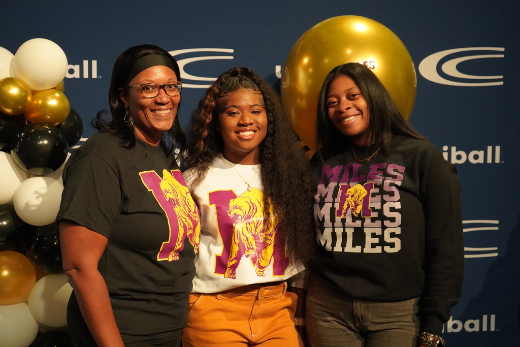 Clay-Chalkville High School student Ky’Morah Bagley smiles while standing next to two adult women. All three of them wear Miles College Sweatshirts and stand in front of a photo backdrop with balloons and Clay-Chalkville's logo on it.