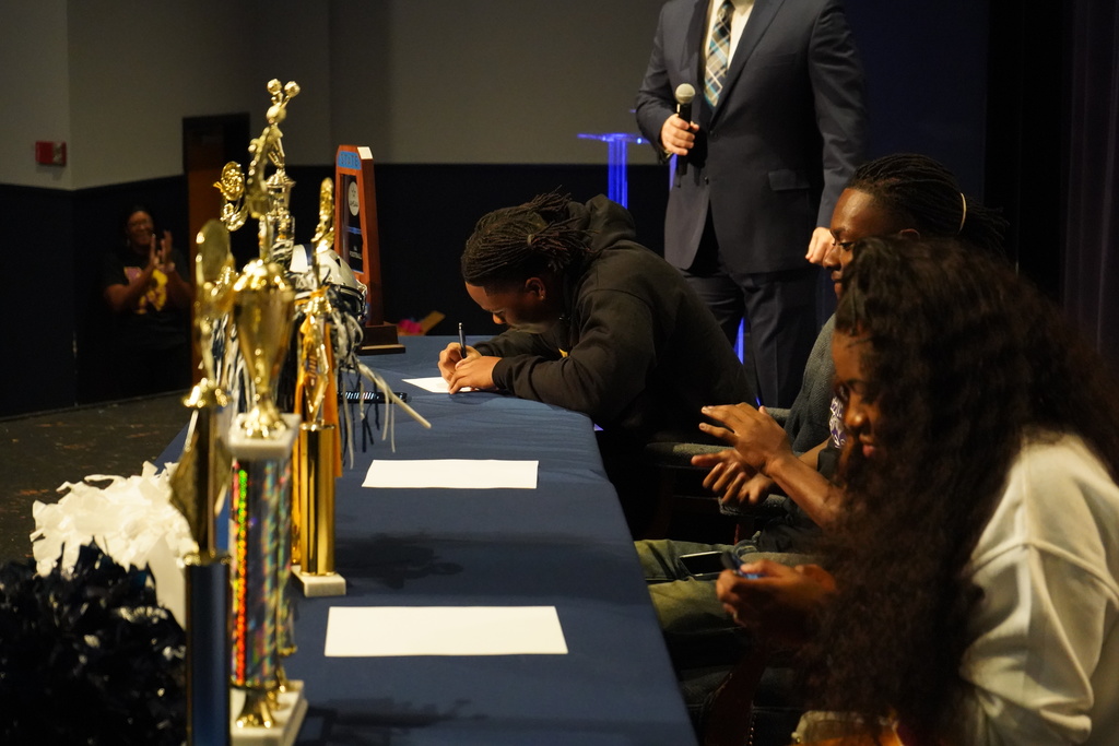 Clay-Chalkville High School student Joshua Woods signs a piece of paper while sitting at a table with two other students during Clay-Chalkville High School's National Signing Day ceremony.