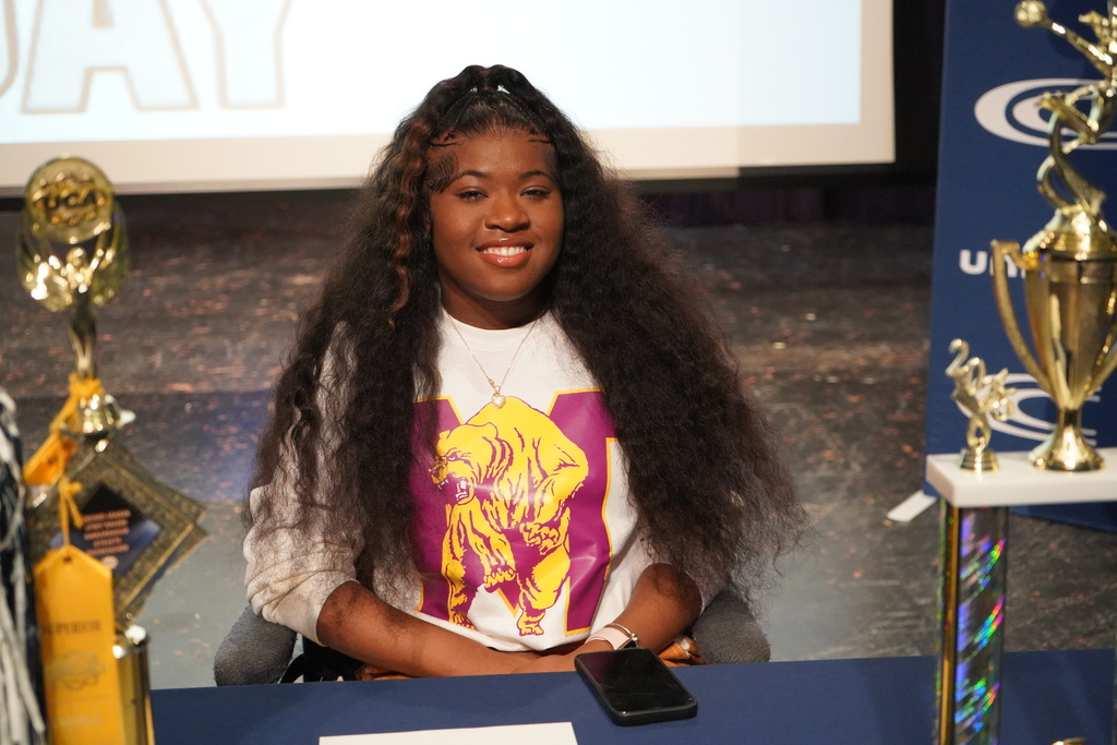 Clay-Chalkville High School student Ky’Morah Bagley smiles while sitting at a table on stage at Clay-Chalkville High School's National Signing Day ceremony.
