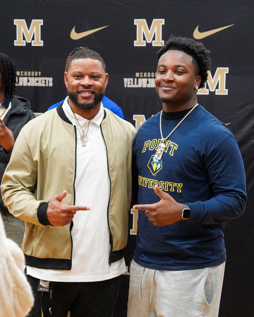 A football coach and a student-athlete pose for a picture on National Signing Day at McAdory High School. They are smiling and pointing at each other. 