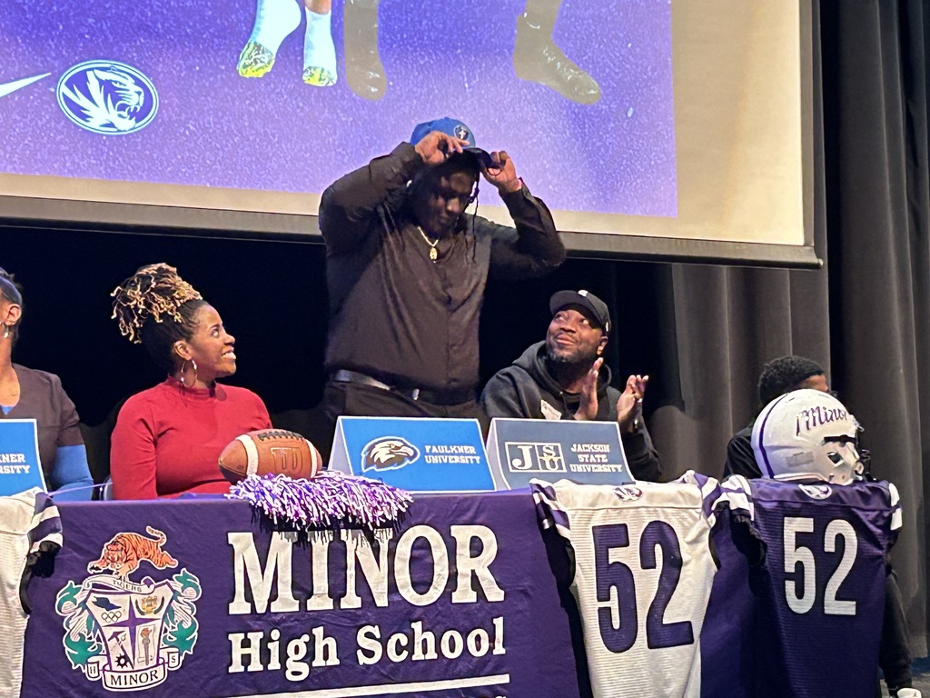 A Minor High School student puts on a blue hat and smiles while standing next to a man and a woman at Minor High School's National Signing Day Ceremony.