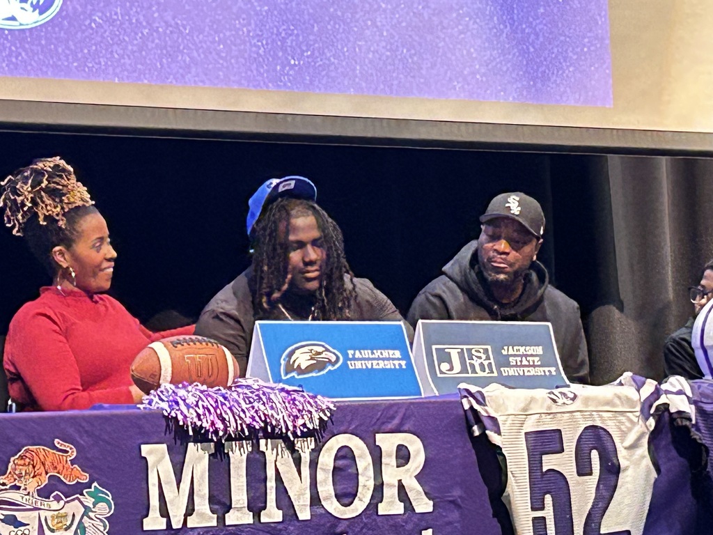 A Minor High School student wears a blue hat and smiles while sitting next to a man and a woman at Minor High School's National Signing Day Ceremony.