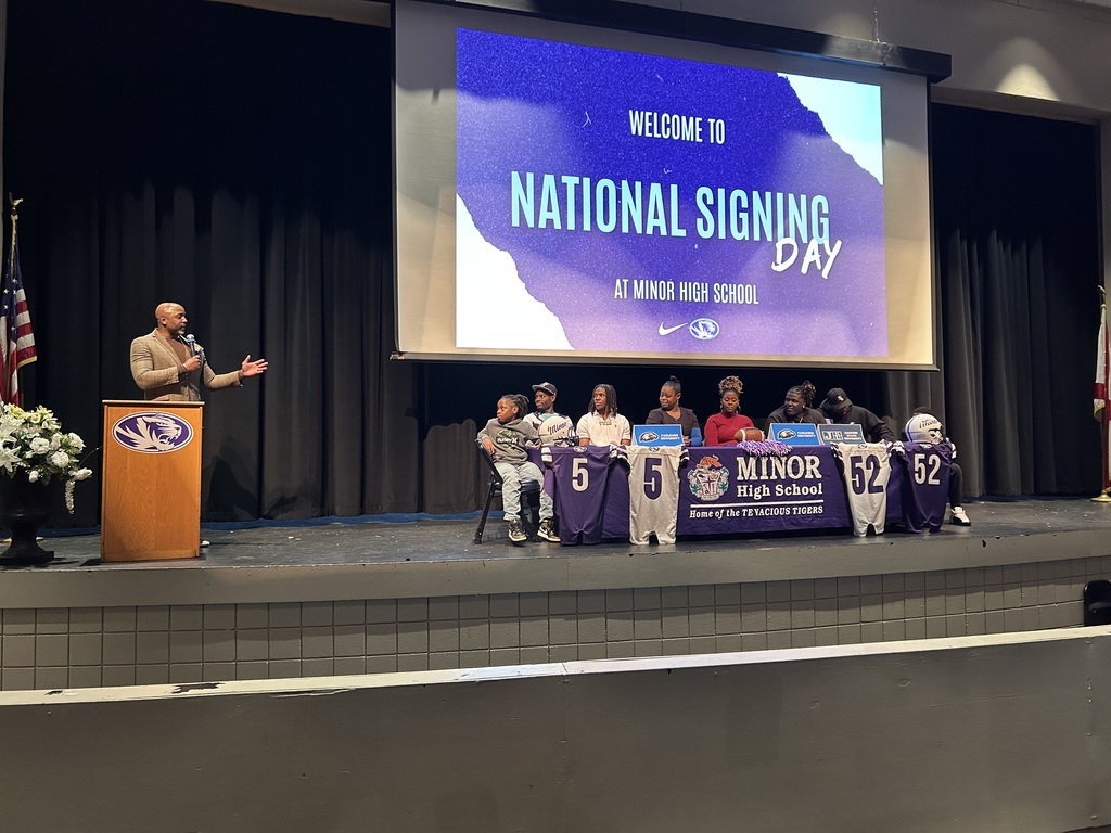 A man speaks into a microphone while men and women sit at a table in Minor High School's Auditorium for the school's National Signing Day Ceremony.